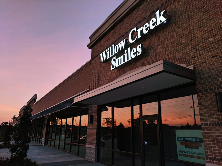 The exterior of a brick commercial building at sunset, displaying "Willow Creek Smiles" signage. Large glass windows reflect nearby trees and power lines, while smaller signage includes "J.A. Greene" with a phone number and URL. The sky features a gradient of pink and purple hues.