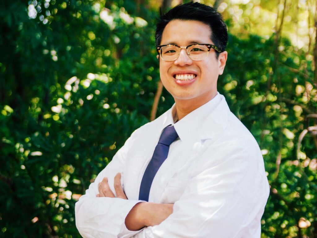 Dr. Nguyen in a white lab coat and dark tie stands with arms crossed in an outdoor setting featuring green foliage.