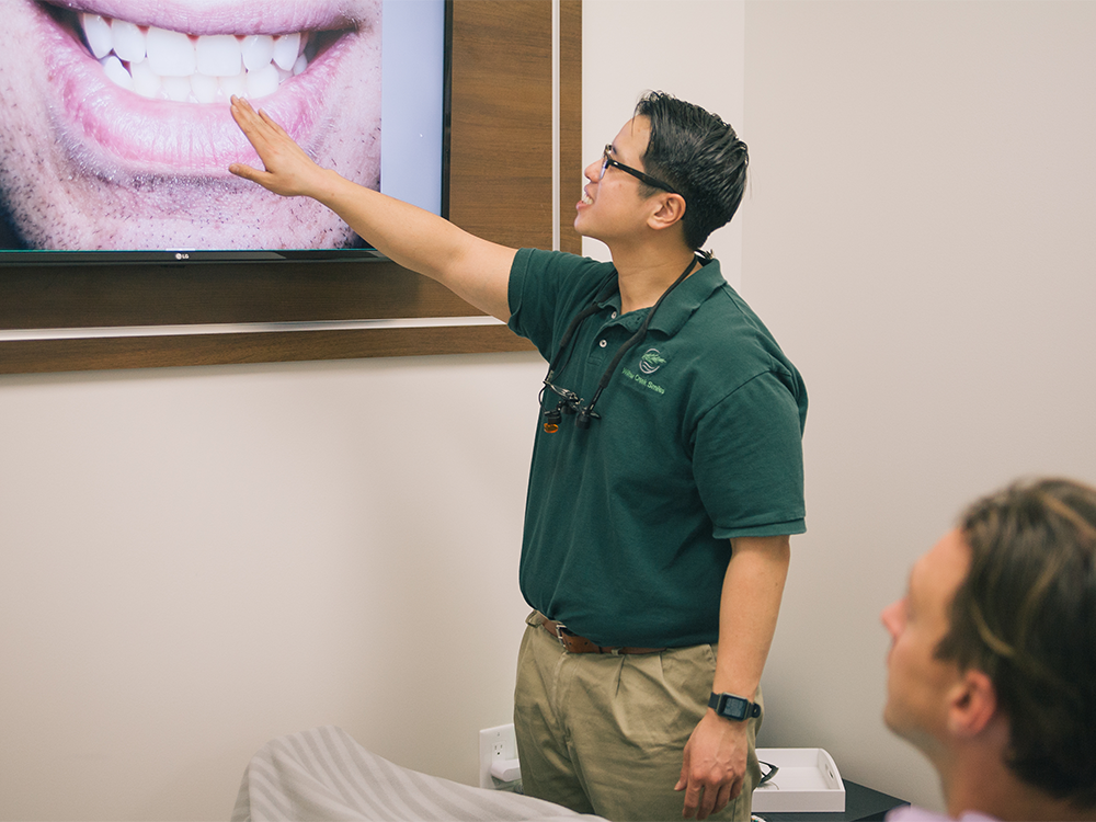 A dental professional in a green polo shirt, with a "Willow Creek Smiles" logo and magnifying glasses, points at a screen showing a close-up image of teeth. In the foreground, part of a seated individual is visible, suggesting a clinical consultation.