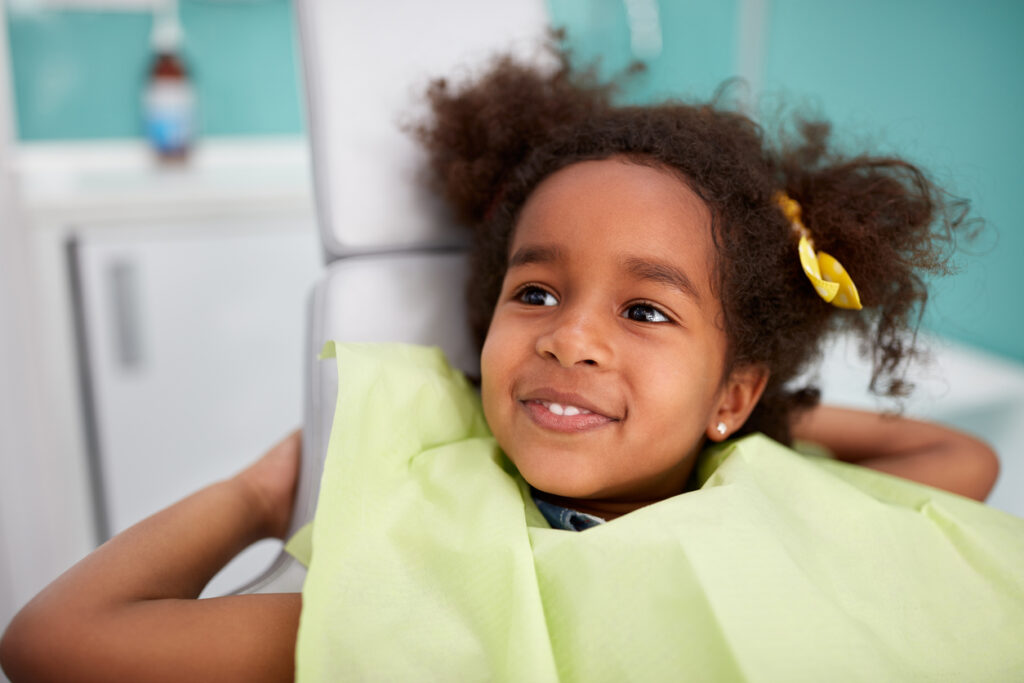 A young girl sits in a dental chair wearing a green bib, with the surrounding objects blurred. Their hairstyle features a yellow hair accessory, and the setting suggests a dental procedure.