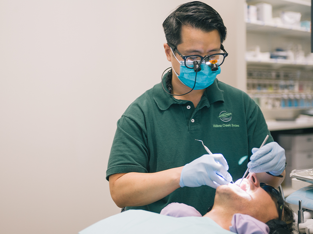 A dental professional in a green polo shirt with "Willow Creek Smiles" embroidered on it performs a procedure. The patient lies back with a protective bib and sunglasses, while dental instruments and supplies are visible in the background.