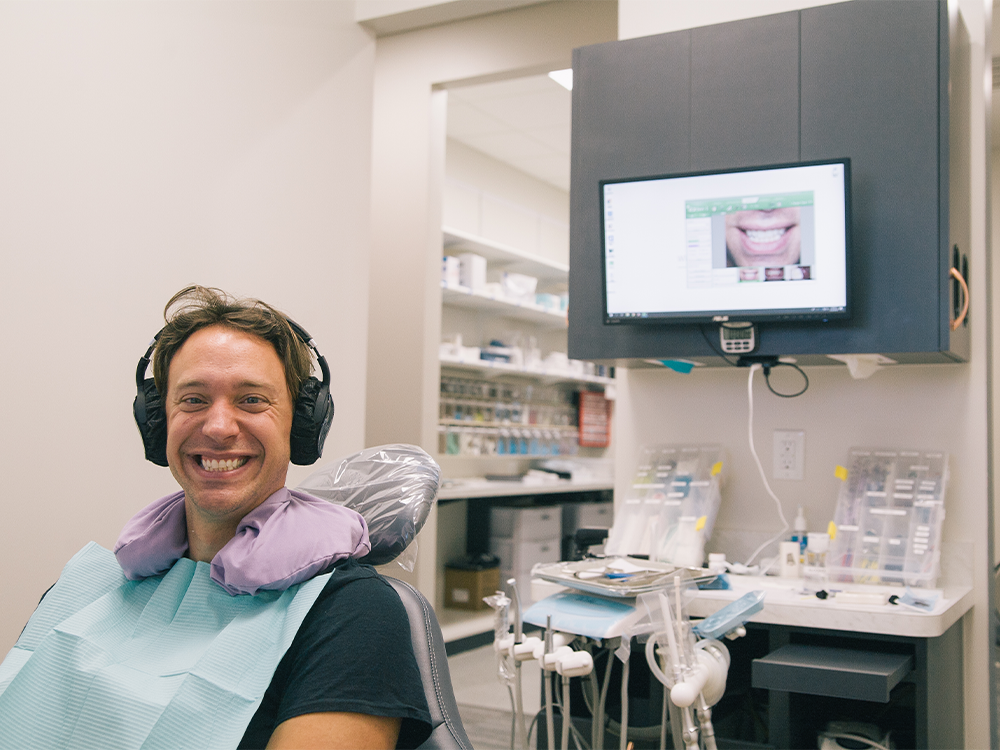 A person sits in a dental office, wearing headphones and a dental bib, positioned in a dental chair. A monitor on the wall displays an image of a smile, while dental tools and equipment are visible in the background.