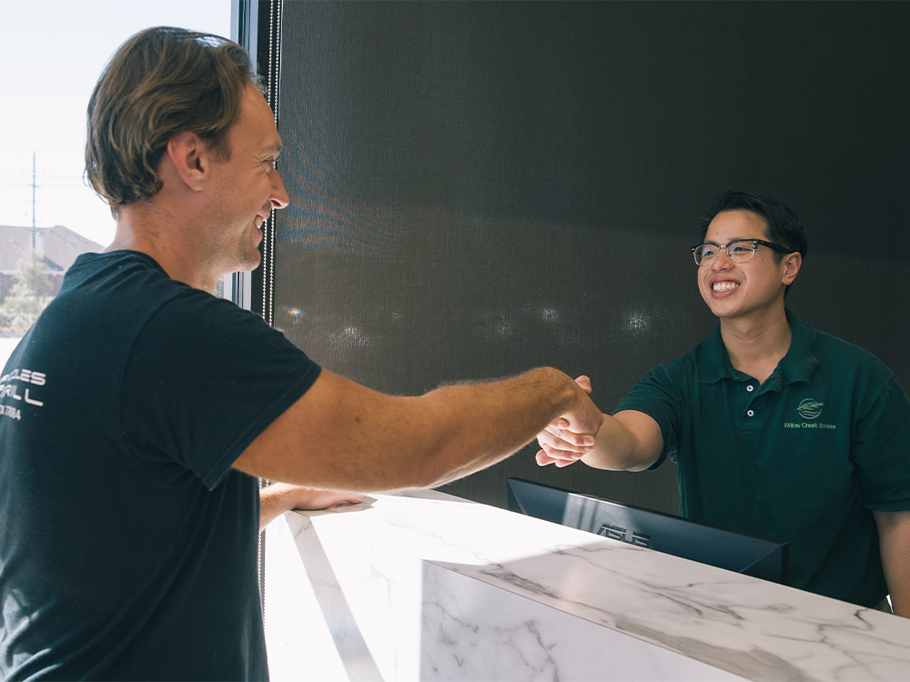 Two individuals shake hands across a marble counter in what appears to be a reception area or service desk. One wears a black t-shirt, while the other dons a dark green polo with a logo reading "Willow Creek Smiles."