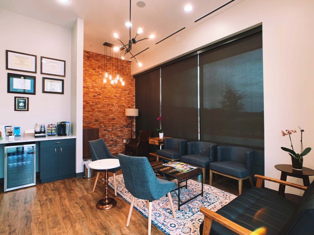 A modern waiting room with a cozy design featuring a black sofa, dark blue chairs, and a small round marble-top table. The space includes framed certificates on the wall, potted plants with colorful flowers, and a refreshment area with a coffee maker. A brick accent wall and hanging light fixtures add warmth to the ambiance.
