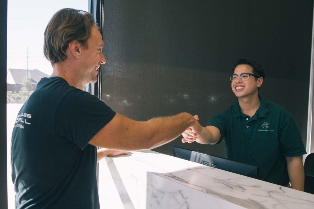Two individuals shake hands across a marble counter in what appears to be a reception area or service desk. One wears a black t-shirt, while the other dons a dark green polo with a logo reading "Willow Creek Smiles."