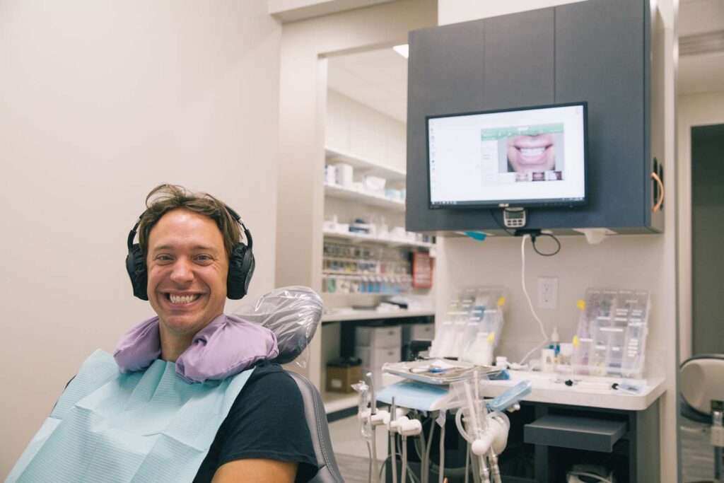 A person sits in a dental office, wearing headphones and a dental bib, positioned in a dental chair. A monitor on the wall displays an image of a smile, while dental tools and equipment are visible in the background.