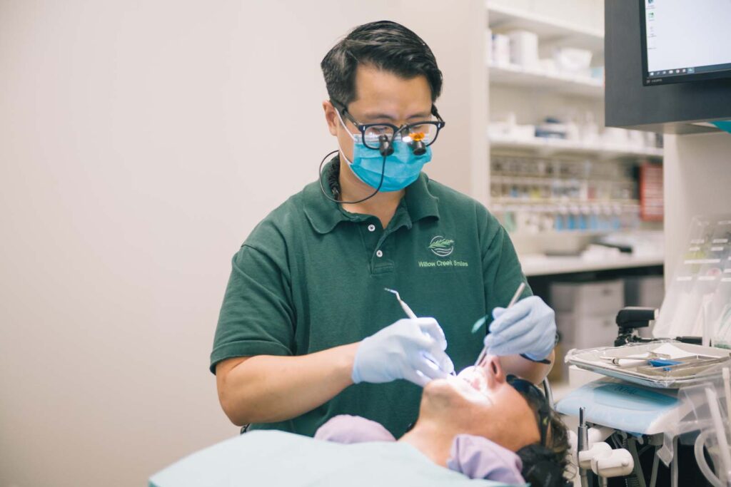 A dental professional in a green polo shirt with "Willow Creek Smiles" embroidered on it performs a procedure. The patient lies back with a protective bib and sunglasses, while dental instruments and supplies are visible in the background.