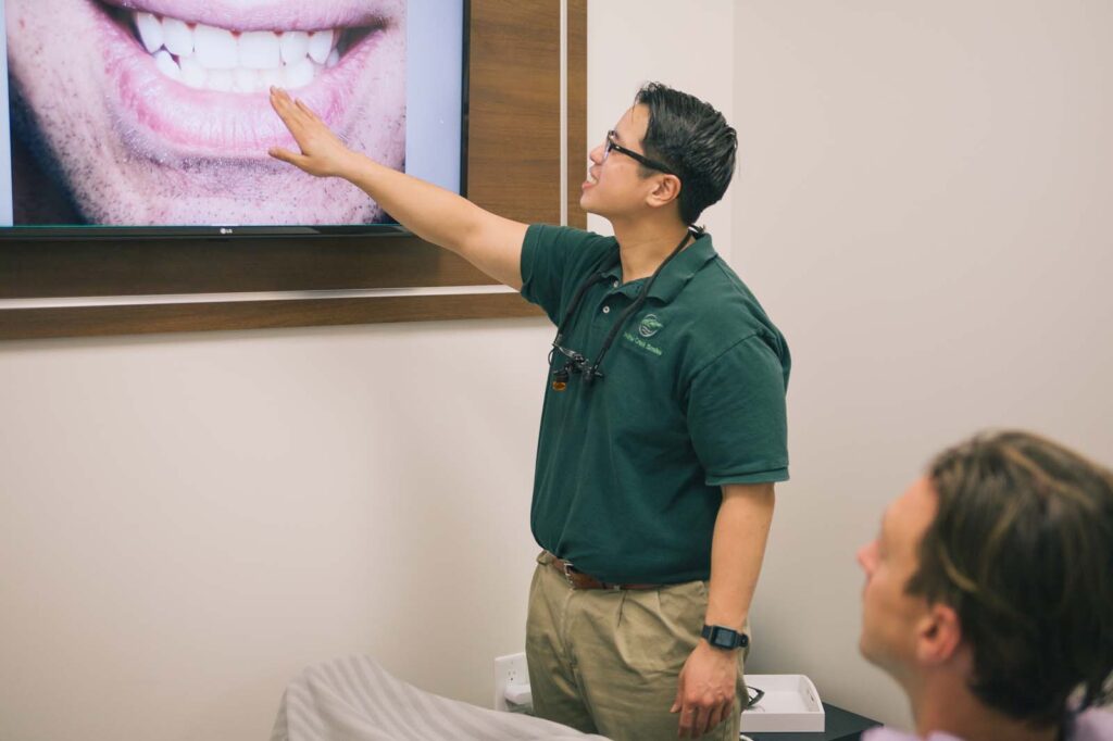 A dental professional in a green polo shirt, with a "Willow Creek Smiles" logo and magnifying glasses, points at a screen showing a close-up image of teeth. In the foreground, part of a seated individual is visible, suggesting a clinical consultation.